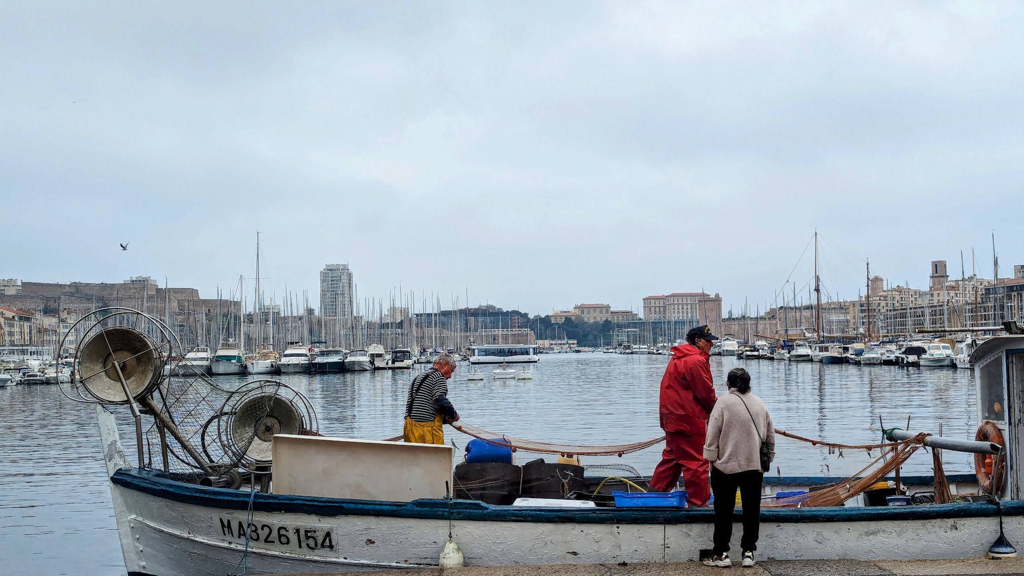 A Picture of Fishermen sorting nets on the port of marseille with the new electric ferry in the backgroun found on our self guided food tour of marseille
