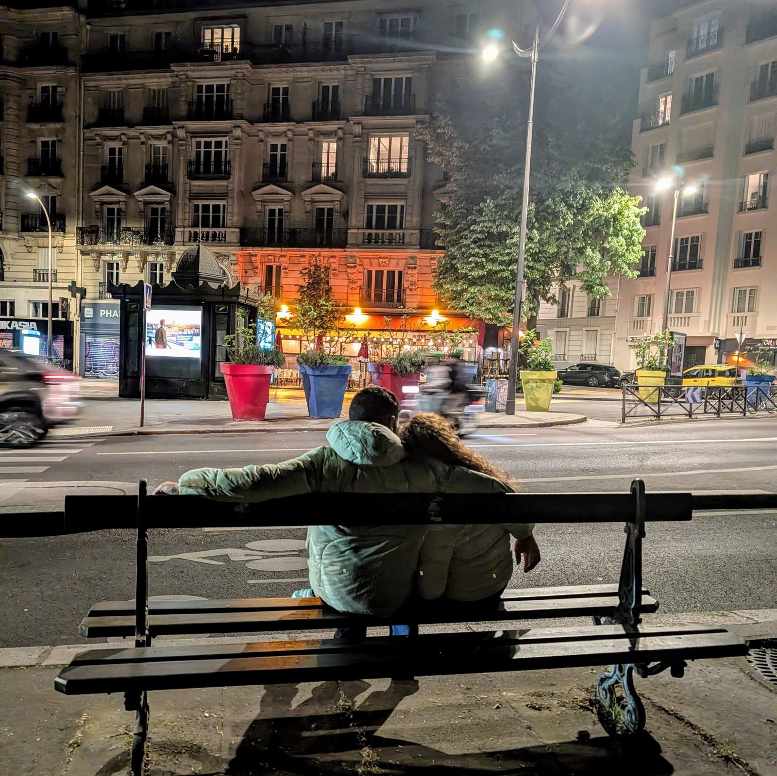 Les Foudres - Romantic couple in front Nighttime view of Les Foudres Paris with glowing terrace lights, as a young couple embraces on a bench across the street.