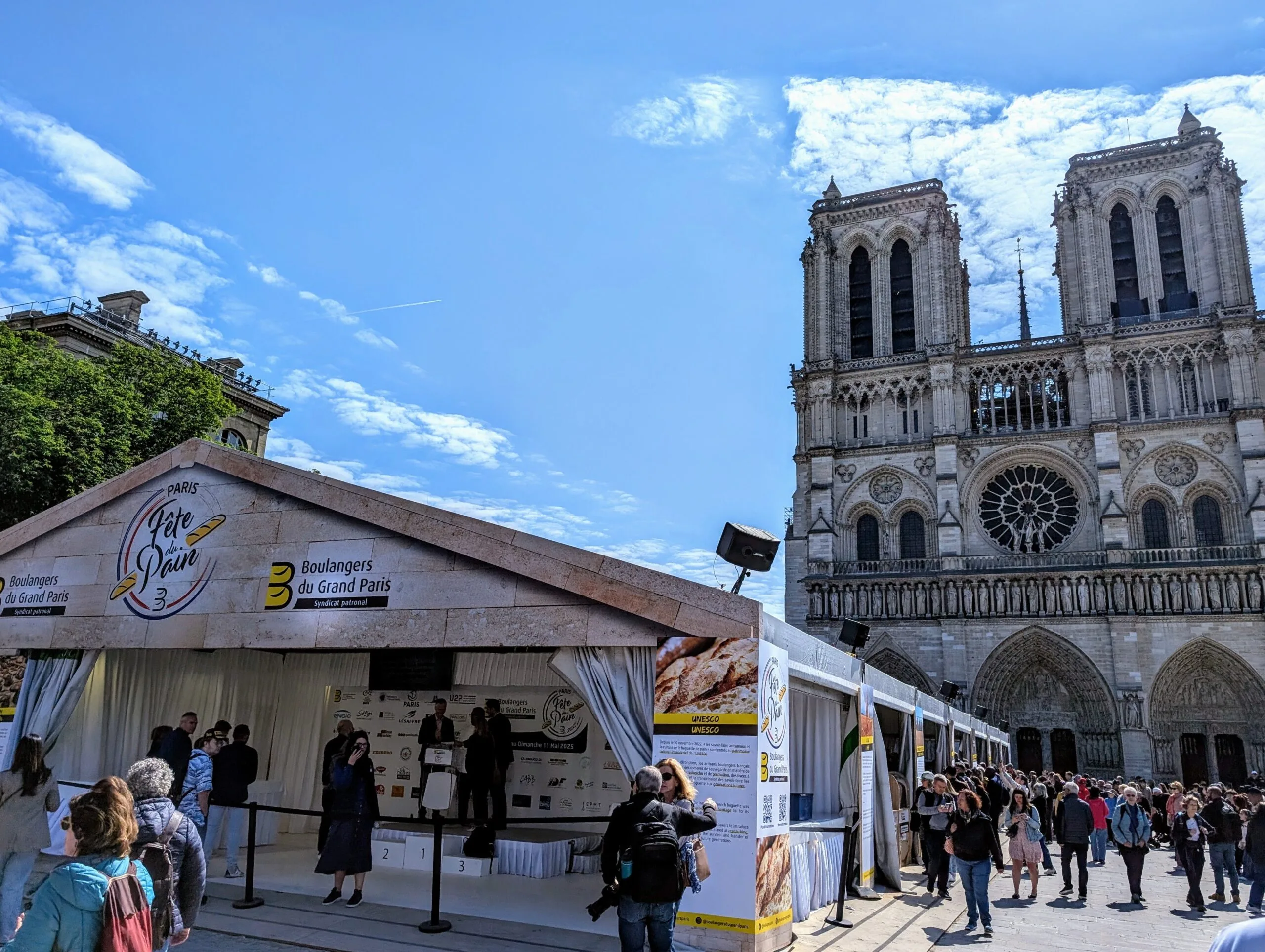 Crowds at the Paris Bread Festival 2025 on the Parvis de Notre-Dame, with bakers on stage beneath the Fête du Pain tent and the cathedral in the background
