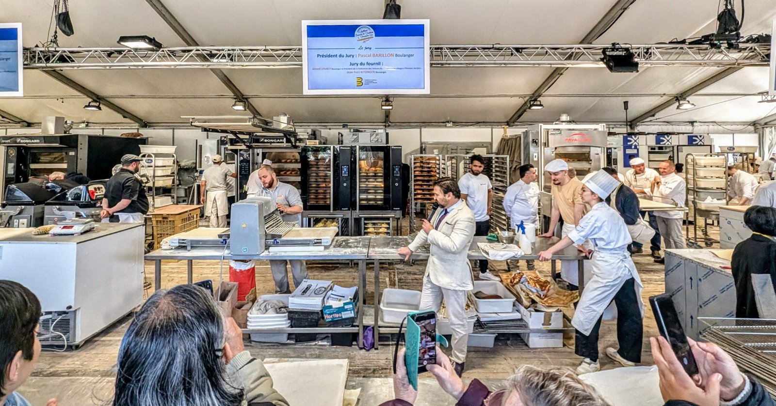 Paris Bread Festival - inside the tent Presenter at Paris Bread Festival 2025 demonstrating the laminator machine while Ferrandi apprentices and French bakers prepare croissants under the festival tent